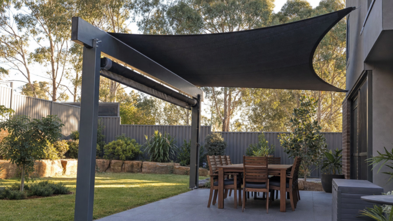 Charcoal shade sail tensioned over an Australian backyard patio with a rolled outdoor blind on the beam and a timber outdoor table in late afternoon light